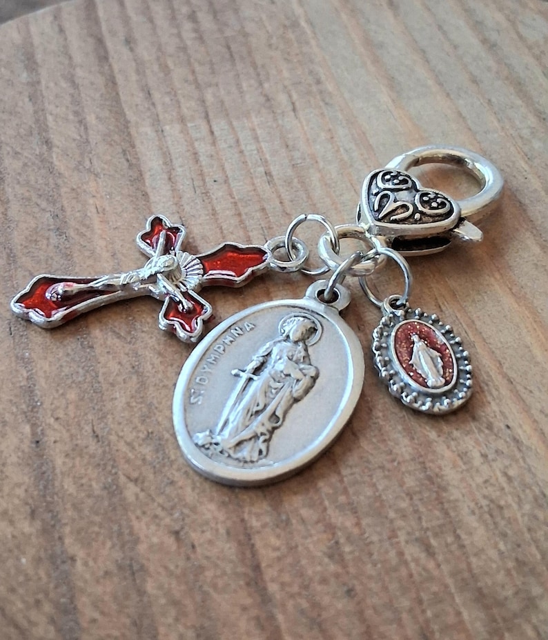 May include: A silver-toned metal charm featuring a heart-shaped lobster clasp attached to three pendants: a red enamel crucifix, an oval medal embossed with the figure of Saint Dymphna, and a smaller oval medal with a red glitter inlay of a figure.