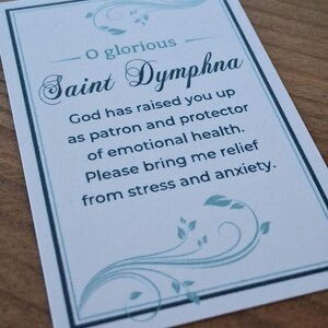 May include: A rectangular prayer card featuring a white background with a light blue border and decorative leaf flourishes. The text reads: O glorious Saint Dymphna, God has raised you up as patron and protector of emotional health. Please bring me relief from stress and anxiety.