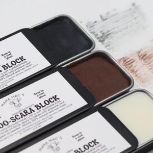 May include: Close-up of three open tins of Mama Mac's Moo-Scara Block. The tins contain black, brown, and white blocks of mascara. The packaging includes text and a cow logo. Swatches of the mascara are visible.