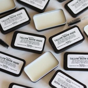 May include: Multiple small black tins with white labels that read "Tallow Brow-Made" and contain a white balm. The balm is used to style eyebrows. The tins are scattered on a white surface with small black brushes.