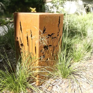 May include: A rusty metal planter with a cutout design of tall grasses and flowers. The planter is sitting on a bed of brown mulch and green grass.