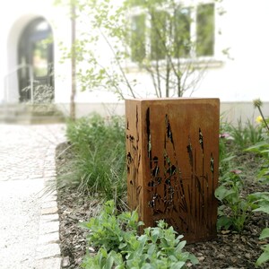 May include: A rusty metal planter with a cutout design of tall grasses. The planter is placed in a garden setting with green plants and brown mulch.