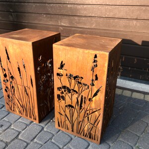 May include: Two rusty metal planters with cut-out designs of tall grasses and flowers. The planters are square and have a rustic, weathered look.