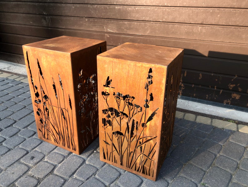 May include: Two rusty metal planters with cut-out designs of tall grasses and flowers. The planters are square and have a rustic, weathered look.