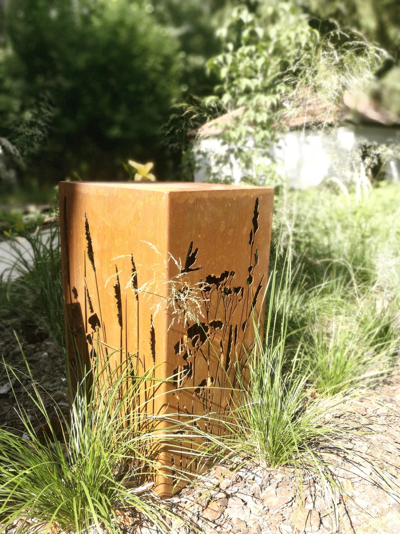 May include: A rusty metal planter with a cutout design of tall grasses and flowers. The planter is sitting on a bed of brown mulch and green grass.