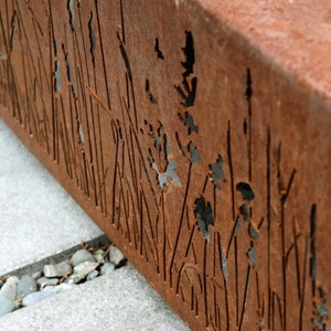 Pu&ograve; includere: Primo piano di un pannello di metallo arrugginito con un design ritagliato di erba alta e fiori. Il pannello &egrave; posizionato su uno sfondo di cemento grigio.