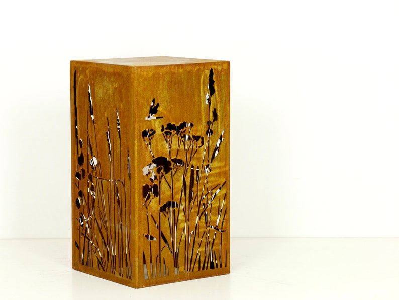 May include: A rusty metal planter with a cutout design of tall grasses and flowers. The planter is square and has a rustic look.