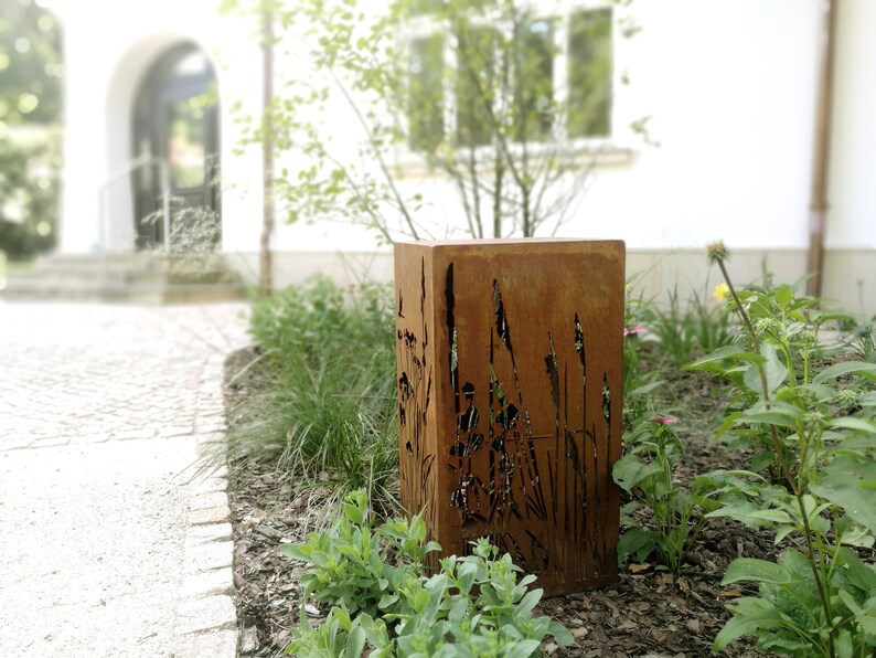 May include: A rusty metal planter with a cutout design of tall grasses. The planter is placed in a garden setting with green plants and brown mulch.
