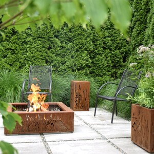Pu&ograve; includere: Un focolare con una cornice in metallo e un design a taglio di erba alta. Il focolare &egrave; circondato da un patio con pav&eacute; grigi. Sullo sfondo ci sono due sedie in metallo nero e una pianta in vaso con foglie verdi.
