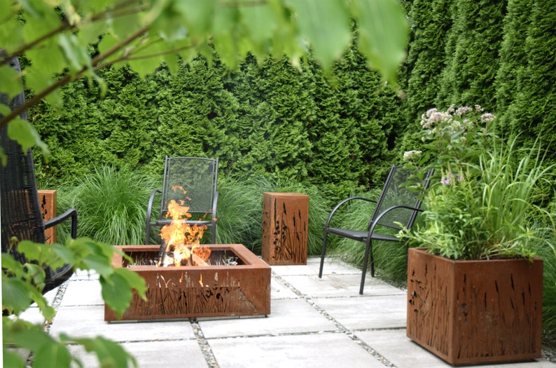 Pu&ograve; includere: Un focolare con una cornice in metallo e un design a taglio di erba alta. Il focolare &egrave; circondato da un patio con pav&eacute; grigi. Sullo sfondo ci sono due sedie in metallo nero e una pianta in vaso con foglie verdi.