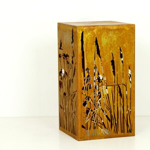 May include: A rusty metal planter with a cut-out design of tall grasses and flowers. The planter is square and has a rustic look.
