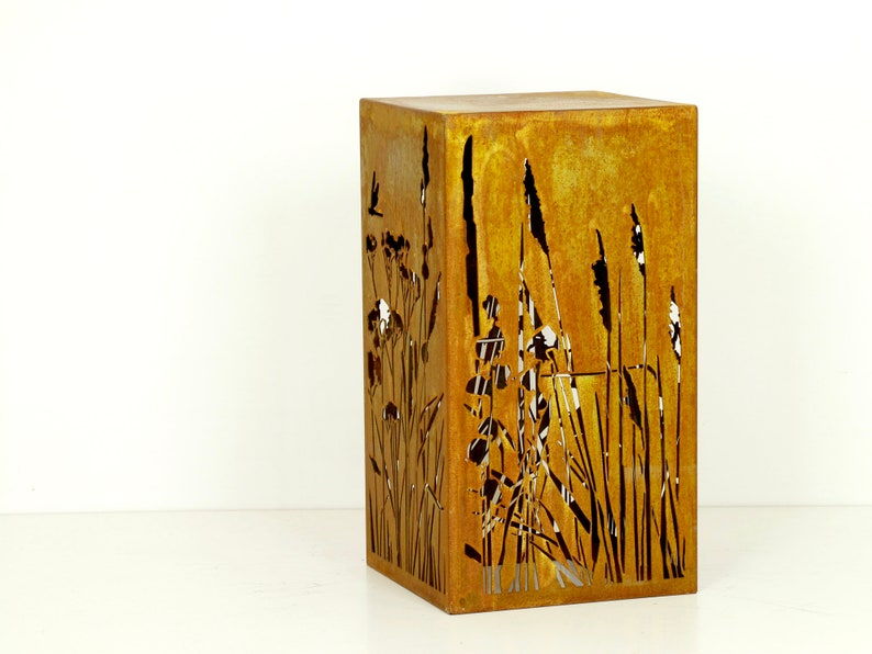 May include: A rusty metal planter with a cut-out design of tall grasses and flowers. The planter is square and has a rustic look.
