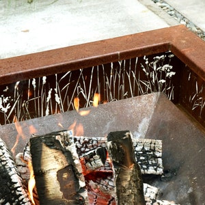 Pu&ograve; includere: Un focolare in metallo arrugginito con un designo decorativo a taglio di erba e fiori bianchi. Il focolare &egrave; pieno di ceppi di legno in fiamme e fiamme.