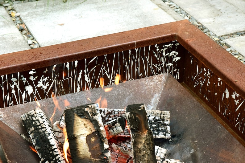 Pu&ograve; includere: Un focolare in metallo arrugginito con un designo decorativo a taglio di erba e fiori bianchi. Il focolare &egrave; pieno di ceppi di legno in fiamme e fiamme.