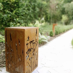 May include: A rusty metal planter with a cutout design of tall grasses and flowers. The planter is sitting on a light-colored stone path.