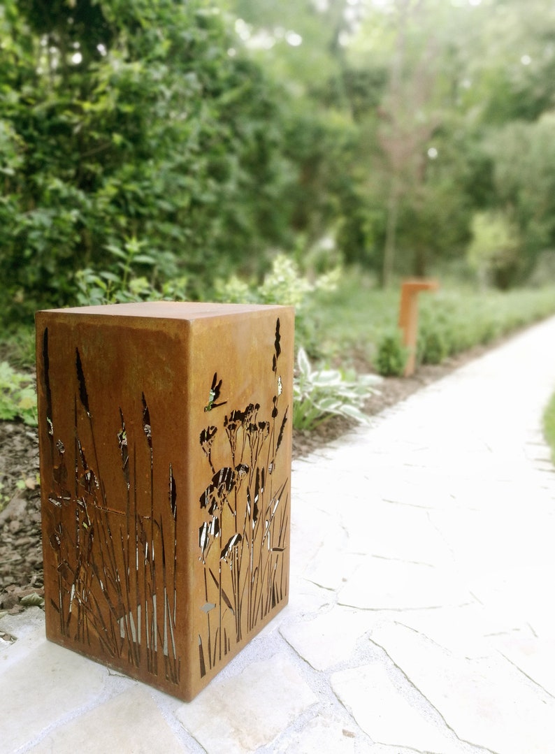 May include: A rusty metal planter with a cutout design of tall grasses and flowers. The planter is sitting on a light-colored stone path.