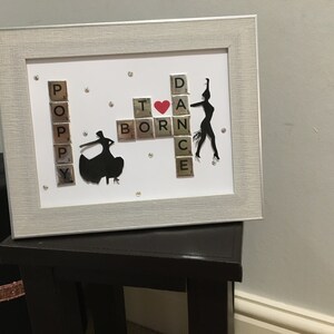 May include: Framed artwork with the words "Poppy Born To Dance" spelled out with Scrabble tiles. Features black silhouette dancers and a red heart. The frame is silver.