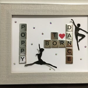 Può includere: Un'opera d'arte incorniciata con sfondo bianco che presenta le parole "Born to Dance" scritte con tessere da Scrabble. La parola "Born" è su una linea orizzontale e la parola "Dance" è su una linea verticale. Un cuore rosso è posizionato tra le parole "Born" e "Dance". Due silhouette nere di ballerini sono su entrambi i lati delle parole. La parola "Poppy" è scritta verticalmente sul lato sinistro dell'opera d'arte.
