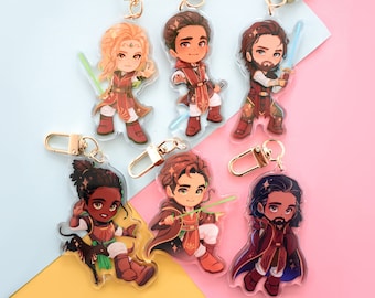 May include: Six colorful character keychains with gold metal rings. Each keychain features a different character with a unique design and pose. The characters are all wearing red and gold clothing and have different hairstyles and facial expressions.
