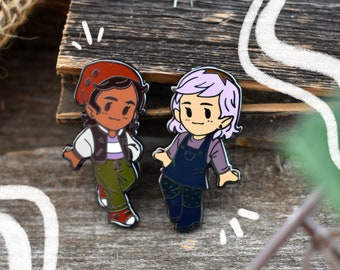 May include: Two enamel pins featuring cartoon characters. The character on the left is wearing a red hat, a white shirt, a green vest, and green pants. The character on the right is wearing a purple shirt and blue overalls. Both characters have brown eyes and are smiling.