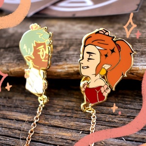 Op de afbeelding: Twee gouden emaillepins met cartoonfiguren met bloemmotieven. De pins zijn verbonden door een gouden ketting. De figuren zijn een persoon met groen haar en een bril en een persoon met rood haar en een rode jurk.