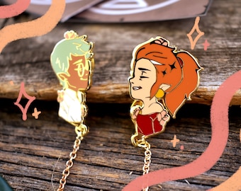 May include: Two gold enamel pins featuring cartoon characters with floral accents. The pins are connected by a gold chain. The characters are a green-haired person with glasses and a red-haired person with a red dress.