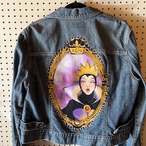 Könnte beinhalten: Eine Jeansjacke mit einem handgemalten Porträt einer Königin in einem goldenen Rahmen auf dem Rücken. Das Porträt hat ein lila und schwarzes Farbschema mit einer goldenen Krone. Die Jacke hat zwei Vordertaschen und einen Knopfverschluss.