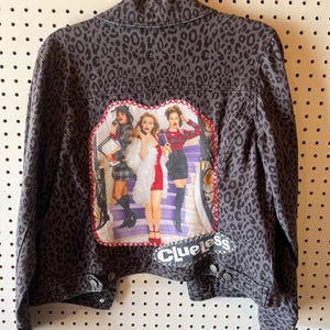 Puede incluir: Chaqueta vaquera gris con estampado de leopardo. La parte trasera presenta un parche en forma de corazón con una imagen colorida de personajes de la película "Clueless" y la palabra "Clueless" en blanco.