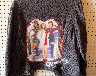 Op de afbeelding: Een grijze spijkerjas met een luipaardprint. De achterkant heeft een hartvormige patch met een kleurrijke afbeelding van personages uit de film "Clueless" en het woord "Clueless" in het wit.