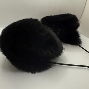 May include: Two black fur earmuffs with long, thin, black stems. The earmuffs are fluffy and round, with a soft texture. The stems are flexible and appear to be made of a rubber-like material. The earmuffs are set against a white background.