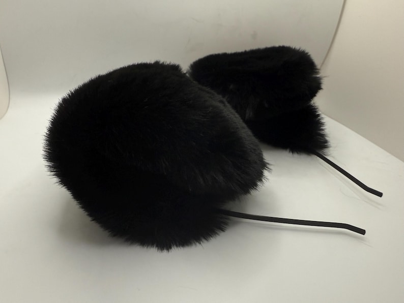 May include: Two black fur earmuffs with long, thin, black stems. The earmuffs are fluffy and round, with a soft texture. The stems are flexible and appear to be made of a rubber-like material. The earmuffs are set against a white background.