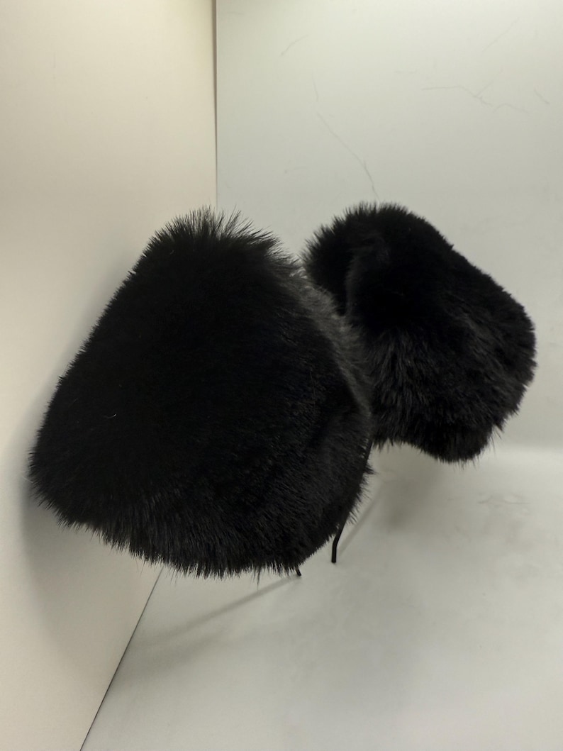 May include: Two black fur earmuffs are displayed against a white background. The earmuffs are fluffy and appear to be made of faux fur. The ear coverings are connected by a thin, black band.
