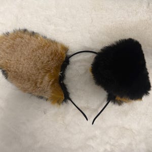Op de afbeelding: Een haarband met twee pluche, imitatiebont dierenoren. Het ene oor is lichtbruin met zwarte bontaccenten, terwijl het andere oor volledig zwart is. De oren zijn bevestigd aan een dunne, flexibele zwarte plastic haarband.