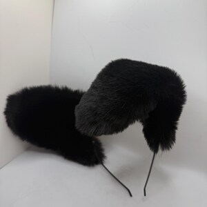May include: A pair of black faux fur cat ears on a thin black headband. The ears are fluffy and upright, with a rounded shape. The headband is made of a thin, flexible wire.