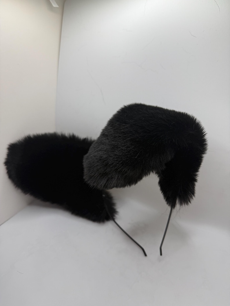 May include: A pair of black faux fur cat ears on a thin black headband. The ears are fluffy and upright, with a rounded shape. The headband is made of a thin, flexible wire.