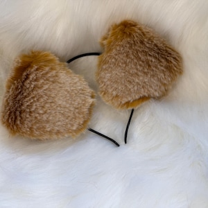 K&ouml;nnte beinhalten: Ein Paar braune, flauschige Ohraccessoires, die an einem schwarzen Stirnband befestigt sind. Die Ohrenformen sind abgerundet und strukturiert und &auml;hneln Tierohren. Der Hintergrund ist ein weiches, wei&szlig;es, pelziges Material.