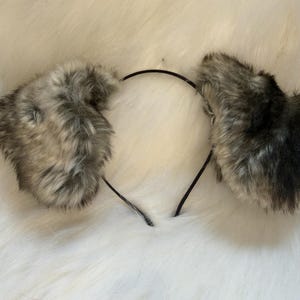 May include: A pair of faux fur animal ears attached to a black headband. The ears are a mix of gray and brown, with a fluffy texture. The headband is thin and flexible, designed for comfortable wear. The ears are likely for cosplay or costume use.