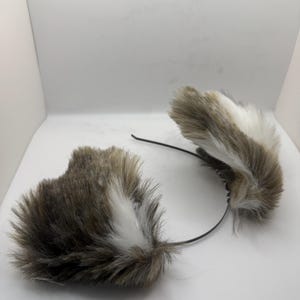 May include: A set of faux fur cat ears on a black headband. The ears are a blend of brown, white, and grey, with a soft, fluffy appearance. The headband is slender and flexible, designed for comfortable wear.