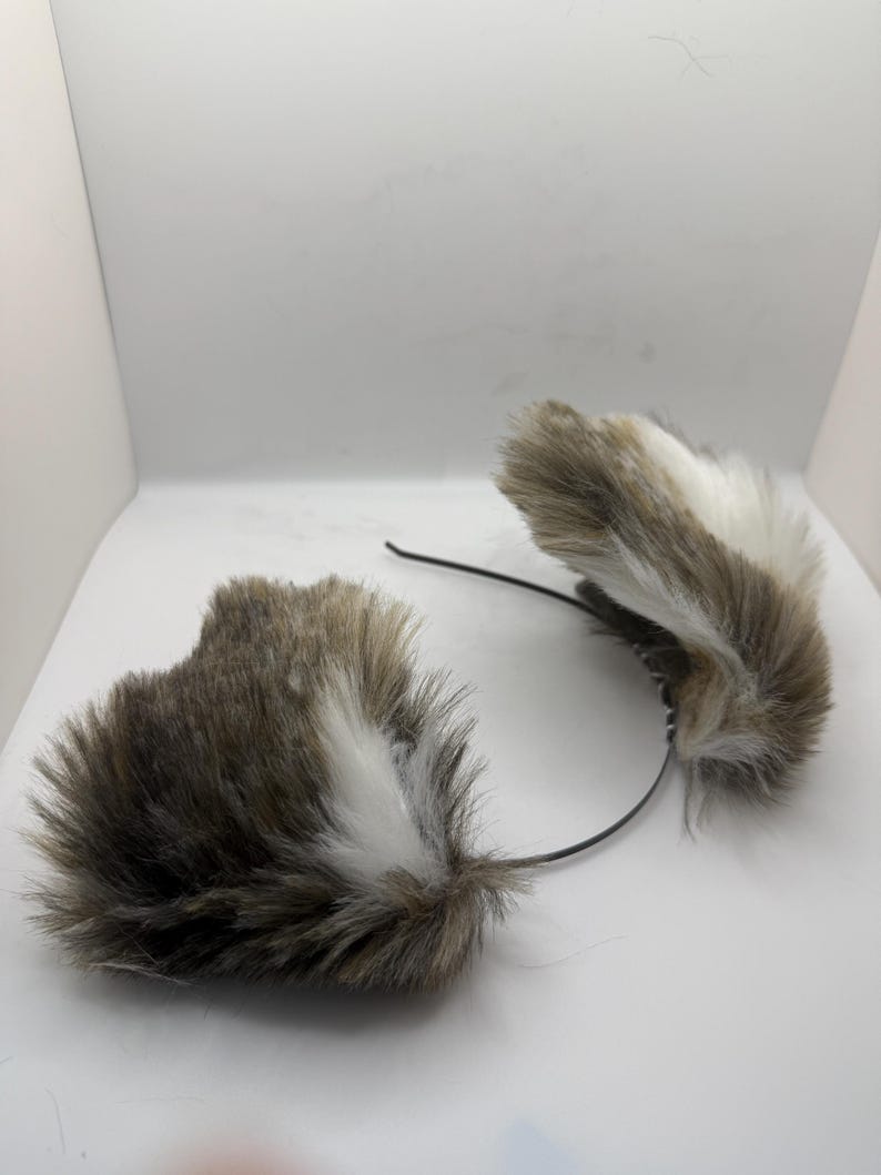 May include: A set of faux fur cat ears on a black headband. The ears are a blend of brown, white, and grey, with a soft, fluffy appearance. The headband is slender and flexible, designed for comfortable wear.