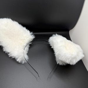 May include: White fluffy cat ears on a black headband. The ears are made of soft, faux fur and are attached to a thin, black metal headband. The accessory is designed to be worn on the head.