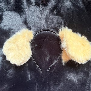 May include: A headband featuring two fluffy, light-yellow dog ears. The ears are made of soft, textured material and are attached to a thin, black headband. This accessory is designed to be worn on the head, suitable for costumes or novelty use.