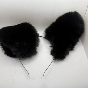 May include: A pair of black faux fur cat ears attached to a thin, black headband. The ears are fluffy and rounded, with a soft texture. The headband is simple and unobtrusive, designed to be worn on the head.