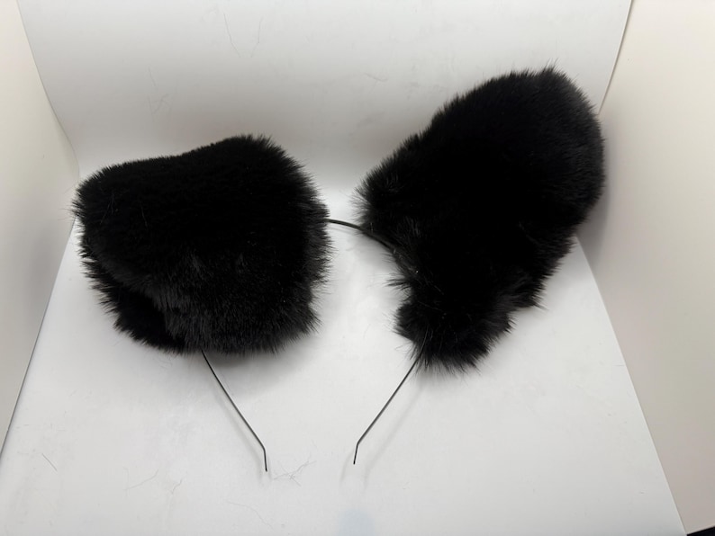 May include: A pair of black faux fur cat ears attached to a thin, black headband. The ears are fluffy and rounded, with a soft texture. The headband is simple and unobtrusive, designed to be worn on the head.