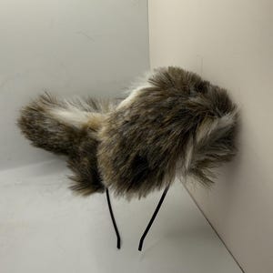 May include: A faux fur headband in shades of brown, white, and black. The headband has a fluffy, textured design and is attached to a thin, black wire frame. This accessory is suitable for costume or fashion.