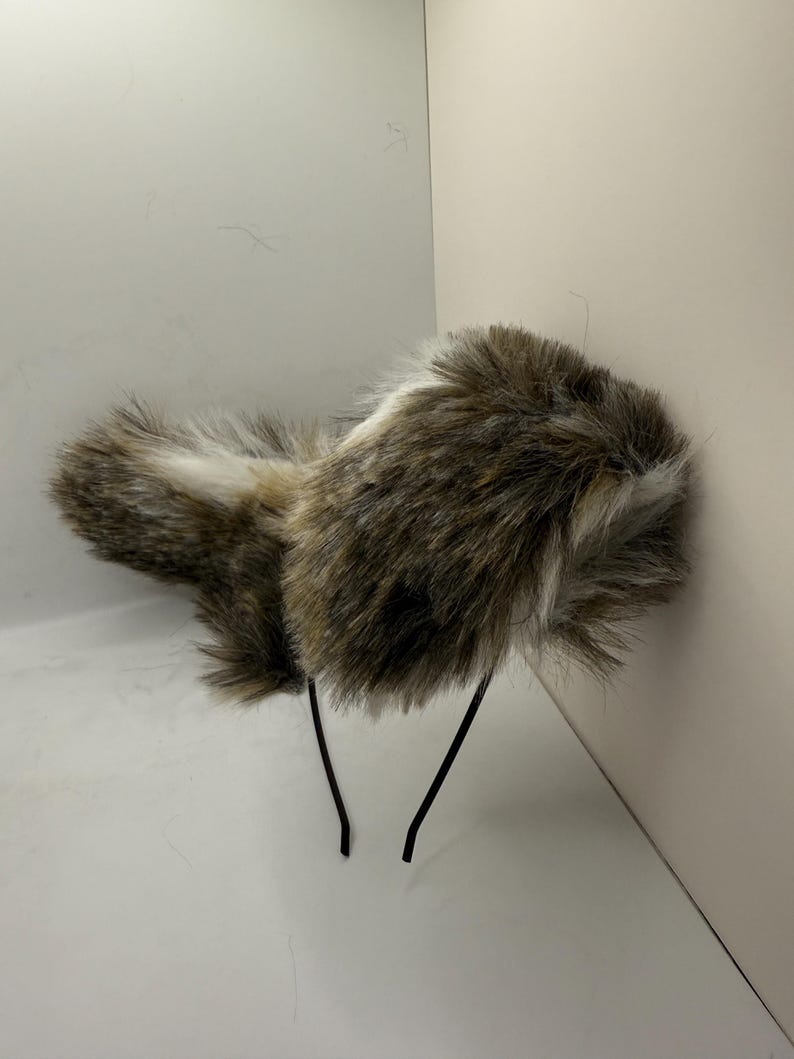 May include: A faux fur headband in shades of brown, white, and black. The headband has a fluffy, textured design and is attached to a thin, black wire frame. This accessory is suitable for costume or fashion.