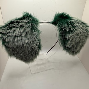 May include: A headband with fluffy, faux fur ear coverings. The ear coverings are a mix of dark green and silver-gray, attached to a thin, black headband. The accessory is displayed on a clear stand against a white background.