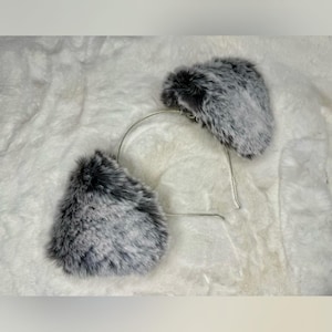May include: A pair of gray and black faux fur cat ears attached to a silver metal headband. The ears are fluffy and textured, with a rounded shape. The headband is thin and flexible, designed to fit comfortably on the head.