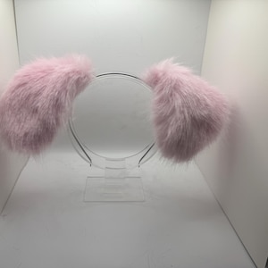 May include: A pair of light pink, fluffy bunny ears attached to a clear plastic headband. The ears are large and rounded, with a soft, furry texture. The headband is displayed on a clear stand.