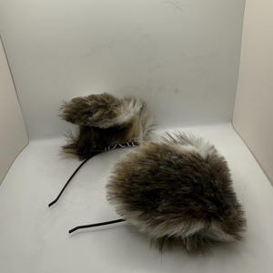 May include: Two faux fur ear headbands. The ear shapes are brown and white, with a fluffy texture. The headbands have a black wire base. The ears are positioned on a white background.