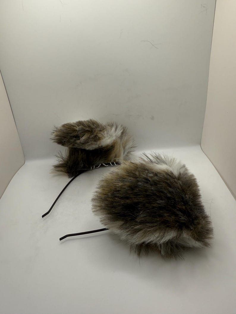 May include: Two faux fur ear headbands. The ear shapes are brown and white, with a fluffy texture. The headbands have a black wire base. The ears are positioned on a white background.